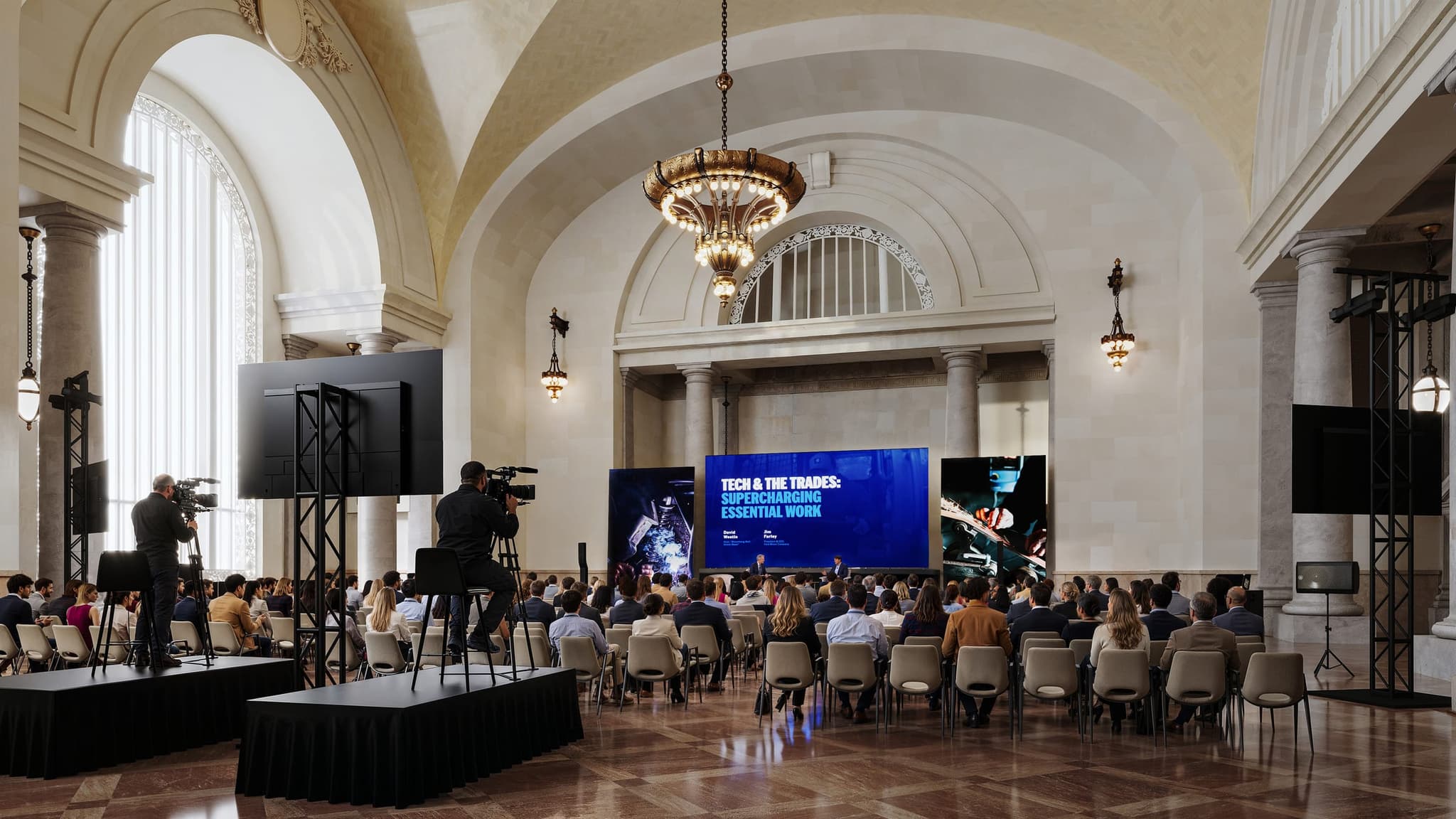 Render of the Grand Hall at Michigan Central Station set for a seated event, with a stage, three screens, camera positions and rows of chairs beneath the vaulted ceiling and chandelier.